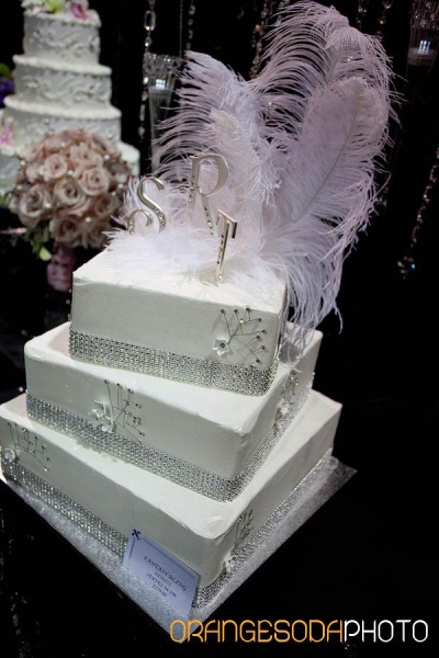 square white cake with sparkling band around each tier, monogram topper with light pink feathers